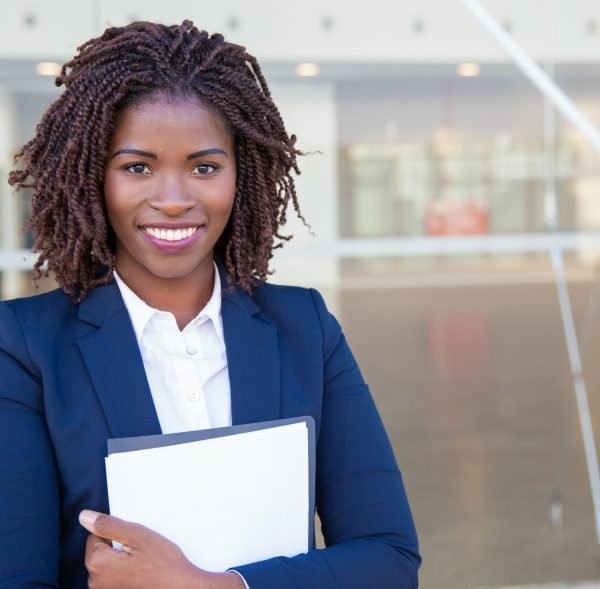 Happy friendly legal expert posing outside. Young black business woman standing at glass wall, holding documents, looking at camera, smiling. Legal expert concept