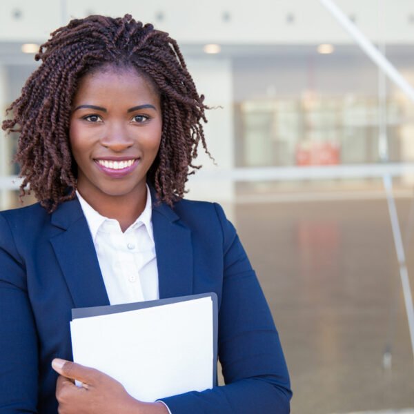 Happy friendly legal expert posing outside. Young black business woman standing at glass wall, holding documents, looking at camera, smiling. Legal expert concept